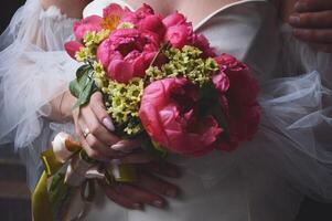 A bride in a white dress with puffy sleeves holds a wedding bouquet of bright pink peonies in her hands. photo