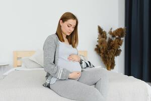 Beautiful pregnant woman holds hands on belly in bedroom at home. Young mother waiting of a baby. Concept of pregnancy, maternity, health care, gynecology, medicine. Close-up, indoors. photo