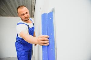 el constructor cuidadosamente corrige el irregularidades de el pared con un paleta. constructor en trabajo ropa en contra un gris pared. foto yesero a trabajar.