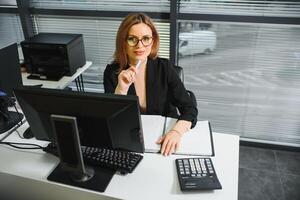 bonito, lindo, lindo, Perfecto mujer sentado a su escritorio en cuero silla en trabajo estación, vistiendo anteojos, Ropa formal, teniendo ordenador portátil y cuaderno en el mesa foto