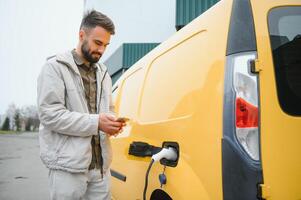 bearded caucasian man standing near an electric car that is charging and making time adjustments on a smartphone. photo