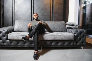 Handsome young african american man in elegant suit with glass of whiskey relaxing on sofa photo