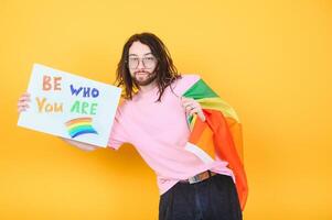 Young queer bisexual smiling gay man with make up in beige tank shirt hold card sign with be who you are title text on rainbow flag background studio portrait. photo