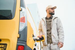 Casual man near electric car waiting for the finish of the battery charging process photo