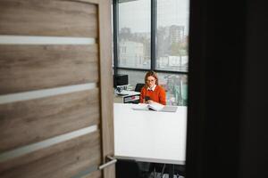 retrato de un contento casual mujer de negocios en suéter sentado a su lugar de trabajo en oficina foto