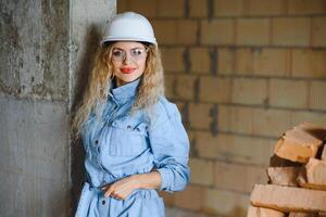 atractivo hembra construcción trabajador en casco de seguridad. confidente joven especialista en a cuadros azul camisa en pantalones en pie en vacío habitación. interior diseño y renovación Servicio foto