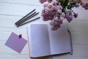 Opened notebook, grey pencils and lilac in vase on a white wooden desktop. photo