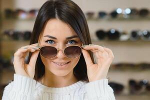 Which one is better fit me. Portrait of cheerful hesitating woman in optician store, making decision, holding stylish sunglasses, choosing what she should buy. Great discounts for buying two pairs. photo