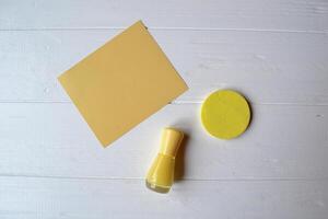 Yellow still life. Female accessories. Reminder on a table. Empty greeting card, nail polish and sponge on a white wooden table. photo