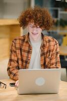 retrato de alegre milenario blogger con moderno Teléfono móvil y ordenador portátil tecnología disfrutando Lanza libre estilo de vida, contento hipster chico en óptico gafas utilizando móvil teléfono y netbook en calle cafetería. foto