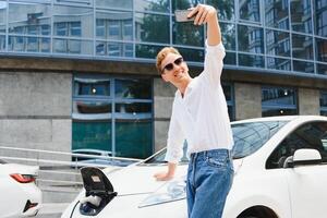 Portrait of young handsome european man, making selfie photo on his smartphone while leaning on his electric car, charging the battery at city power station after buying frsh products in trade mall