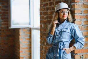 Attractive female construction worker in hardhat. Confident young specialist in checkered blue shirt in jeans standing in empty room. Interior design and renovation service. photo