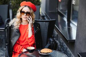 joven elegante mujer en rojo boina teniendo un francés desayuno con café y cuerno sentado exteriores a el café terraza foto