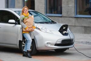 Woman with a mobile phone near recharging electric car. Vehicle charging at public charging station outdoors. Car sharing concept photo