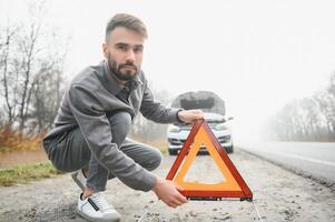 un joven hombre cerca un roto coche con un abierto capucha en el borde del camino. foto