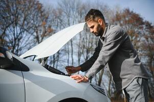 A young man near a broken car with an open hood on the roadside. photo