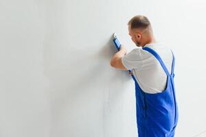 Portrait of a worker in overalls and holding a putty knife in his hands against the plastered wall background. Repair work and construction concept photo