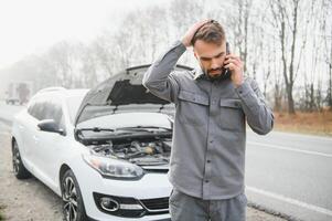 un joven hombre cerca un roto coche con un abierto capucha en el borde del camino. foto