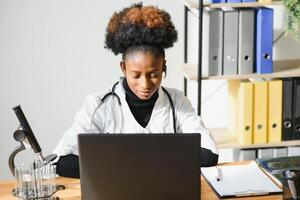 Smiling african american female doctor gp wears white medical coat using laptop computer at workplace gives remote online consultation, working on pc, consulting patient in internet telemedicine chat photo