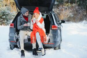 Happy Young couple is sitting in the back trunk of car in winter forest and drinking hot tea from thermos. Winter time photo