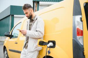 bearded caucasian man standing near an electric car that is charging and making time adjustments on a smartphone. photo