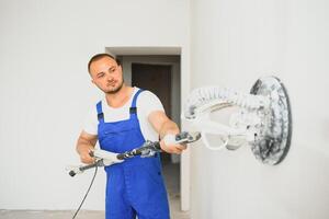 yesero suaviza el pared superficie con un pared amoladora. Maestro constructor moler un blanco yeso pared. un hombre en mono muele el superficie en un respirador. experimentado reparador foto