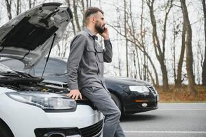 hombre utilizar un Teléfono móvil llamada garaje en frente de el abierto capucha de un roto coche en el la carretera en el bosque. coche Descompostura concepto. foto