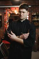 retrato de hermoso joven hombre en pie a Barbero tienda. elegante estilista en pie en su salón con su brazos cruzado. foto