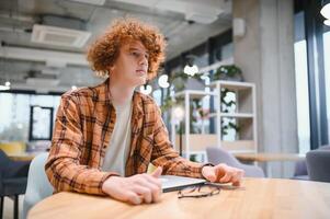 Young happy male freelancer in casual clothes sitting in cafe with laptop and using mobile phone. photo