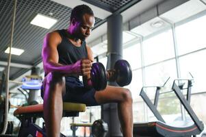 joven africano americano hombre sentado y levantamiento un pesa cerca a el estante a gimnasio foto