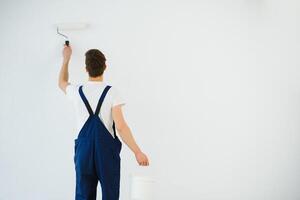 young man in blue overalls painting wall into white color with a roller photo