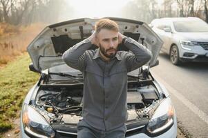 Young upset casual man trying to fix his broken car outdoors. Man waiting for towing service for help car accident on the road. Roadside assistance concept. photo