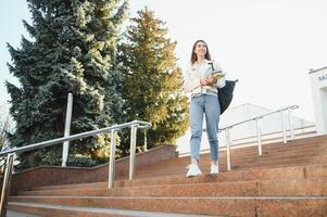 joven niña estudiante sonriente en contra universidad. linda niña estudiante sostiene carpetas y cuadernos en manos. aprendiendo, educación concepto foto