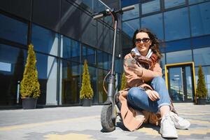 joven hermosa mujer en un chaqueta sonrisas y paseos un eléctrico scooter a trabajo a lo largo oficina edificios foto