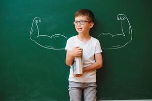 linda niño chico en colegio uniforme y lentes. Vamos a colegio para el primero tiempo. niño con colegio bolso y libros. niño en clase habitación cerca pizarra con músculos en él. espalda a colegio foto