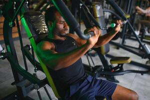 atlético joven negro hombre teniendo rutina de ejercicio en cofre prensa máquina en gimnasio foto