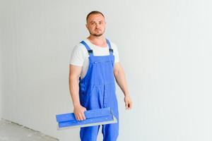 Portrait of a worker in overalls and holding a putty knife in his hands against the plastered wall background. Repair work and construction concept photo