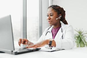 African American woman doctor working at her office online using portable inormation device. Telemedicine services. Primary care consultations, psychotherapy, emergency services photo