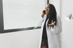 medicamento, tecnología y cuidado de la salud concepto - contento sonriente africano americano hembra médico o en blanco Saco con estetoscopio vocación en teléfono inteligente terminado hospital antecedentes. foto