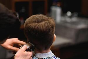 un pequeño linda chico se sienta en un peluquería a el estilista, un alumno es consiguiendo pelo cortar en un belleza salón, un niño a un barberías, un corto de los hombres Corte de pelo foto