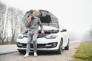 hombre utilizar un Teléfono móvil llamada garaje en frente de el abierto capucha de un roto coche en el la carretera en el bosque. coche Descompostura concepto. foto