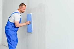 Portrait of a worker in overalls and holding a putty knife in his hands against the plastered wall background. Repair work and construction concept photo