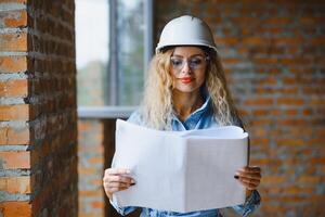 atractivo hembra construcción trabajador en casco de seguridad. confidente joven especialista en a cuadros azul camisa en pantalones en pie en vacío habitación. interior diseño y renovación servicio. foto