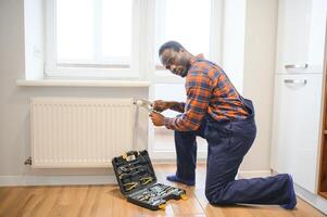 hombre en ropa de trabajo mono utilizando herramientas mientras instalando o reparando calefacción radiador en habitación foto