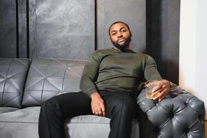 Handsome young african american man in elegant suit with glass of whiskey relaxing on sofa photo