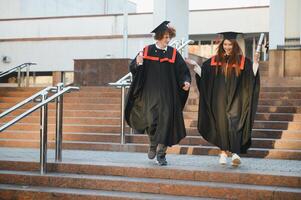 retrato de contento graduados dos amigos en graduación tapas y vestidos en pie fuera de Universidad edificio con otro estudiantes en fondo, participación diploma pergaminos, y sonriente foto