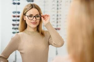 Pretty young woman is choosing new glasses at optics store. photo