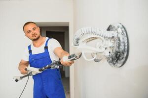 yesero suaviza el pared superficie con un pared amoladora. Maestro constructor moler un blanco yeso pared. un hombre en mono muele el superficie en un respirador. experimentado reparador foto