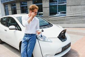 joven hermoso hombre participación cargando cable a eléctrico cargando estación punto en pie cerca su nuevo coche , mirando satisfecho. foto