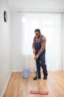 profesional limpiador en azul uniforme Lavado piso y limpiando polvo desde el mueble en el vivo habitación de el departamento. limpieza Servicio concepto foto